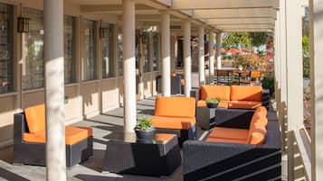 An outdoor seating area with wicker furniture and orange cushions, featuring a glass wall and columns.