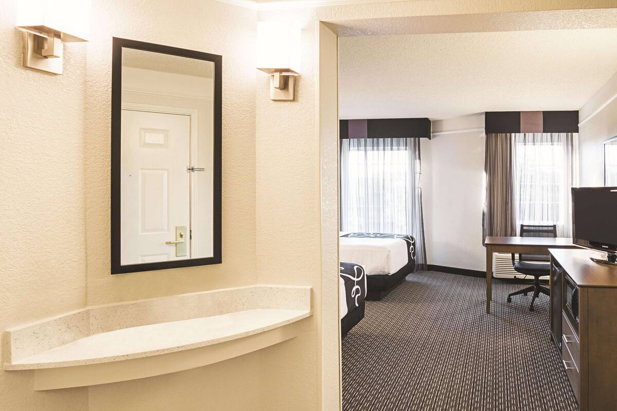 A hotel room with a large mirror, a small sink, two beds, a desk, and a television.