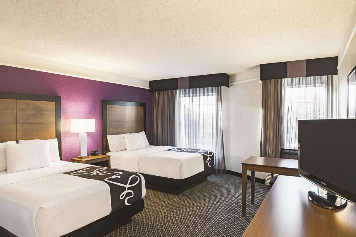 A hotel room with two beds, a desk, a TV, and a window with curtains.