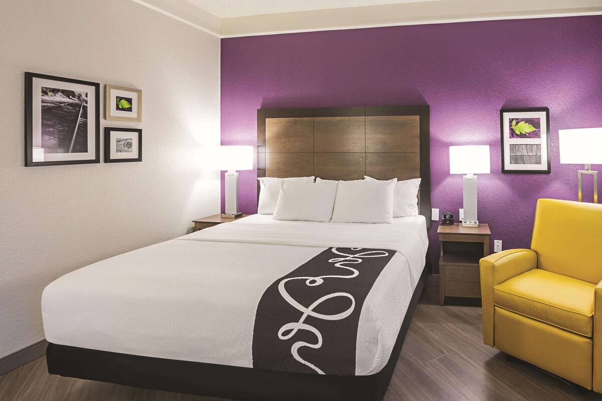 A hotel room with a bed, a nightstand, a chair, and two wall-mounted lamps.