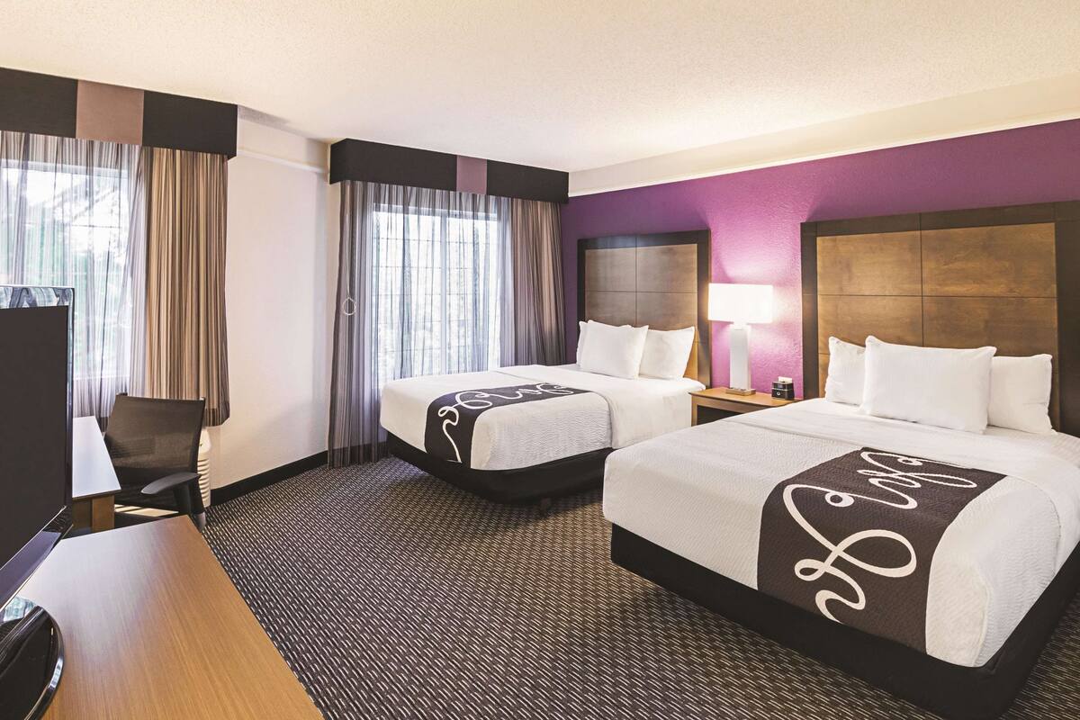 A hotel room with two beds, a desk, a chair, a lamp, and a view of the city through large windows.