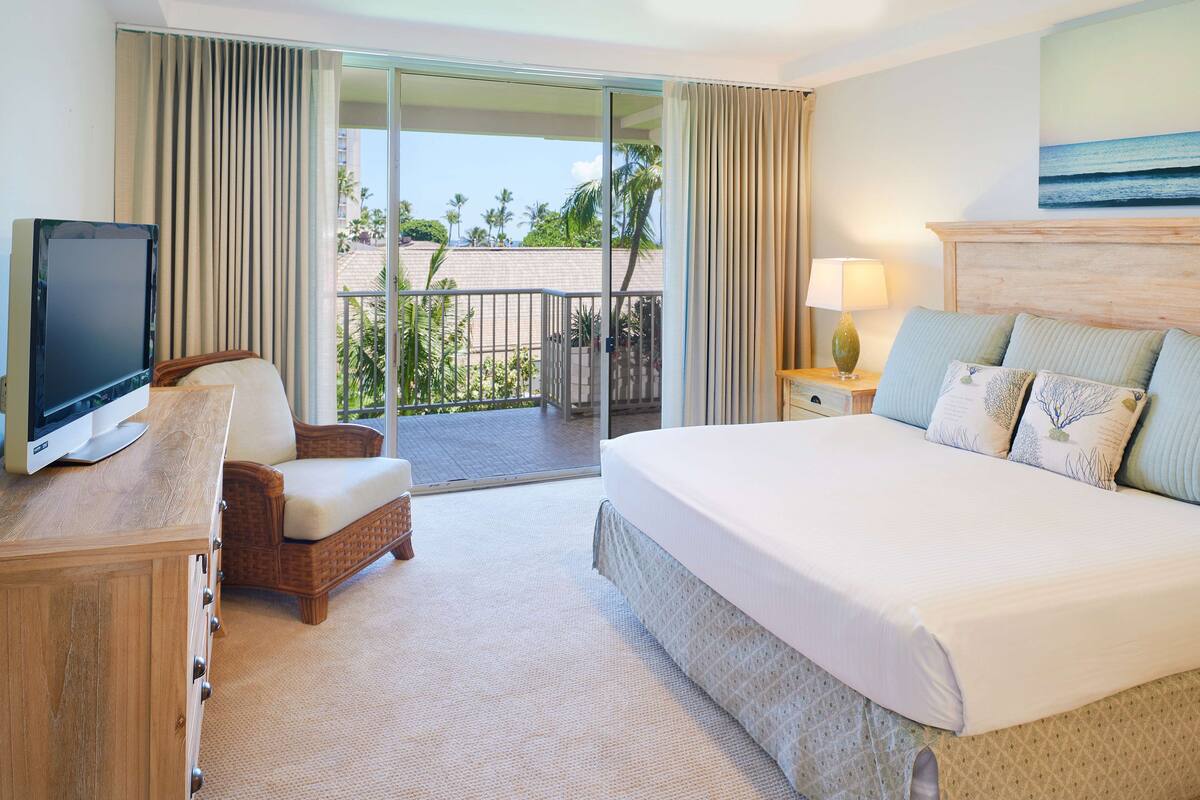 A hotel room with a bed, a TV, a chair, a nightstand, and a balcony with a view of palm trees.