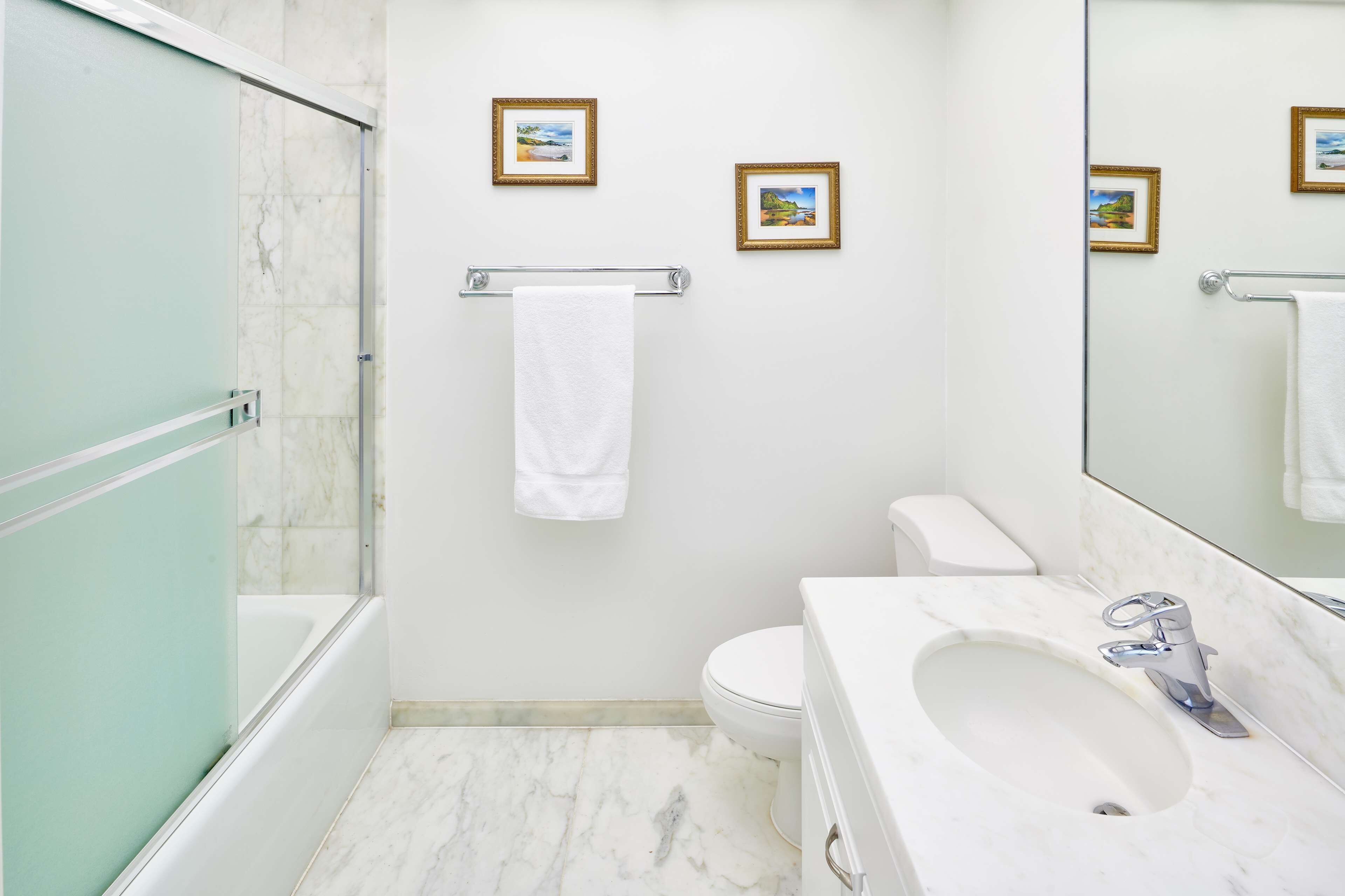 A bathroom with a glass shower enclosure, a toilet, a sink with a mirror, and three framed pictures on the wall.