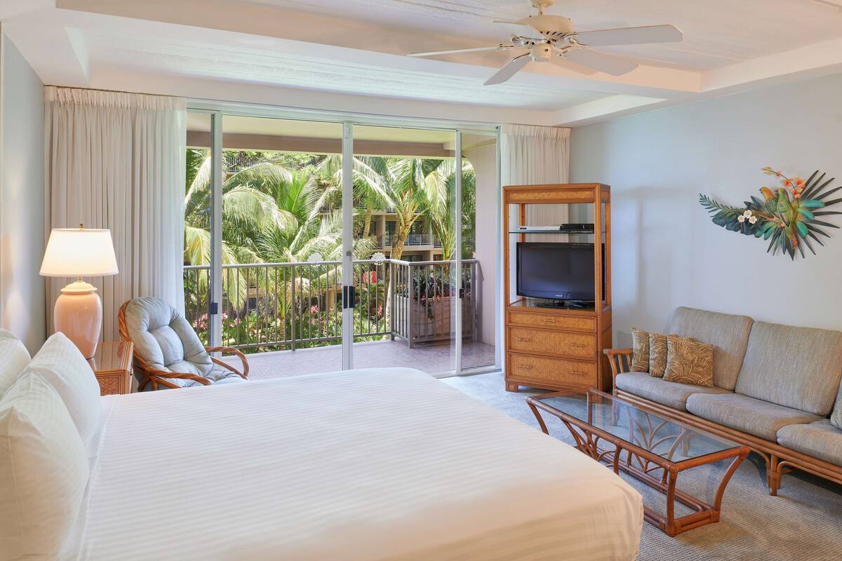 A hotel room with a large bed, a sofa, a TV stand, and a balcony with a view of palm trees.