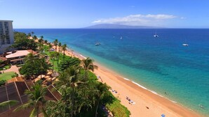 A beach with clear turquoise water, a sandy shore, and several boats. There are palm trees and a building in the background.