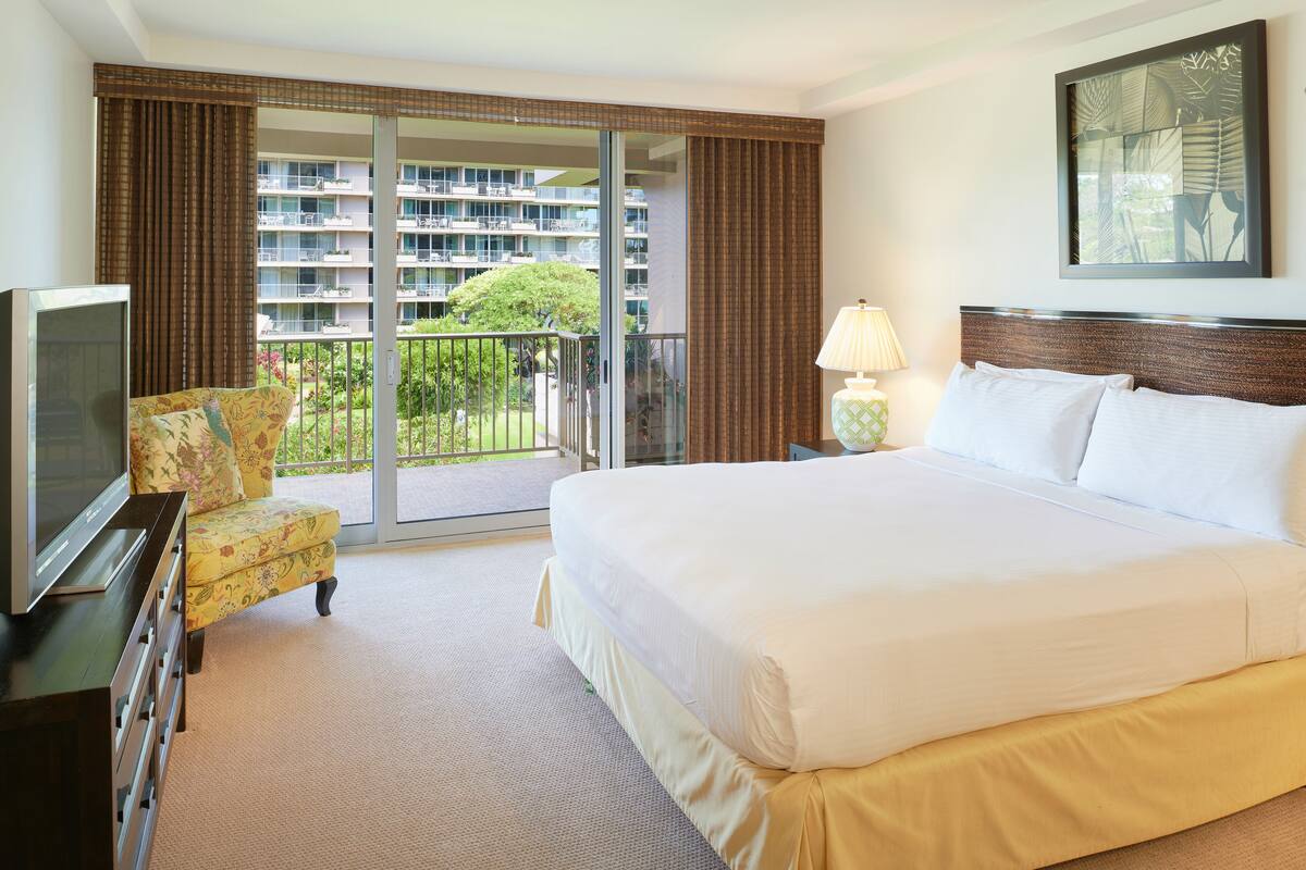 A hotel room with a large bed, a TV, a sofa, and a balcony with a view of a building and greenery.