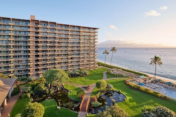 A beach with clear turquoise water, a sandy shore, and several boats. There are palm trees and a building in the background. at Aston at The Whaler on Kaanapali Beach