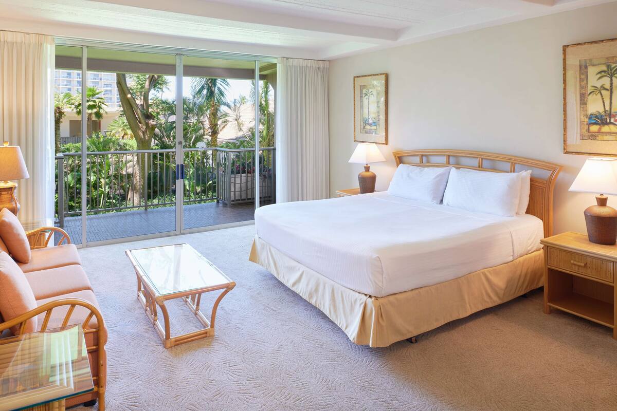 A hotel room with a large bed, a glass coffee table, a sofa, and a balcony with a view of palm trees.