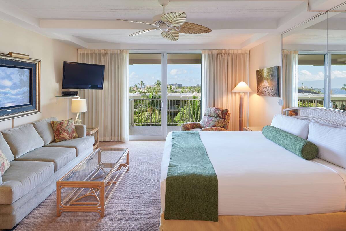 A hotel room with a large bed, a sofa, a TV, and a balcony with a view of palm trees and buildings.