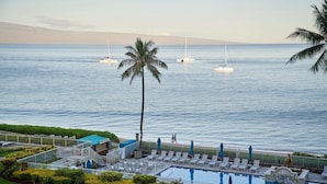 Ein Hotelpool mit Liegestühlen und Sonnenschirmen, eine Palme und Segelboote im Hintergrund.