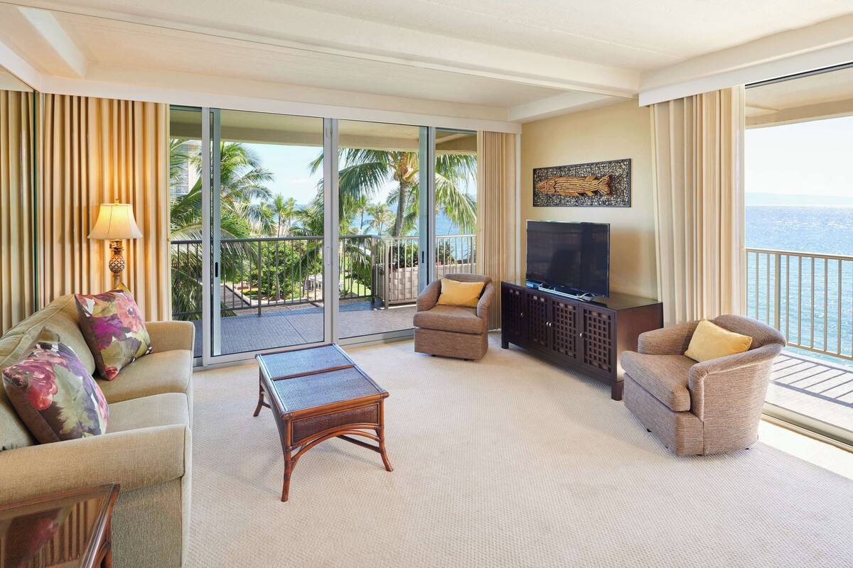 A hotel room with a sofa, armchairs, a coffee table, a TV, and a balcony with a view of palm trees and the ocean.