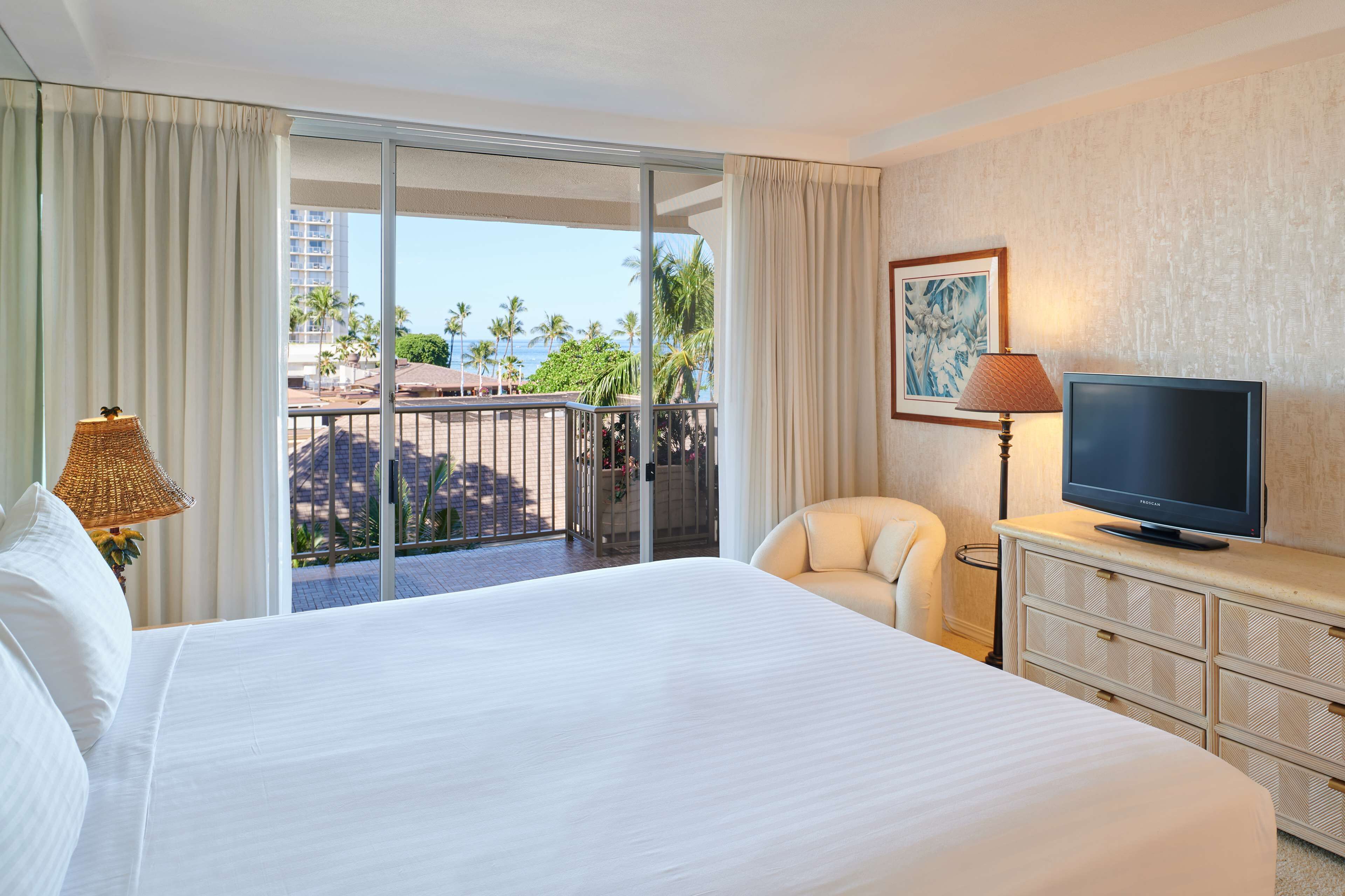 Habitación de hotel con una cama grande, televisión, una silla, cómoda y balcón con vistas a palmeras y edificios.