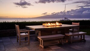 An outdoor dining area with a stone table and benches, overlooking a body of water and a sailboat. - Aston at The Whaler on Kaanapali Beach (Lahaina)