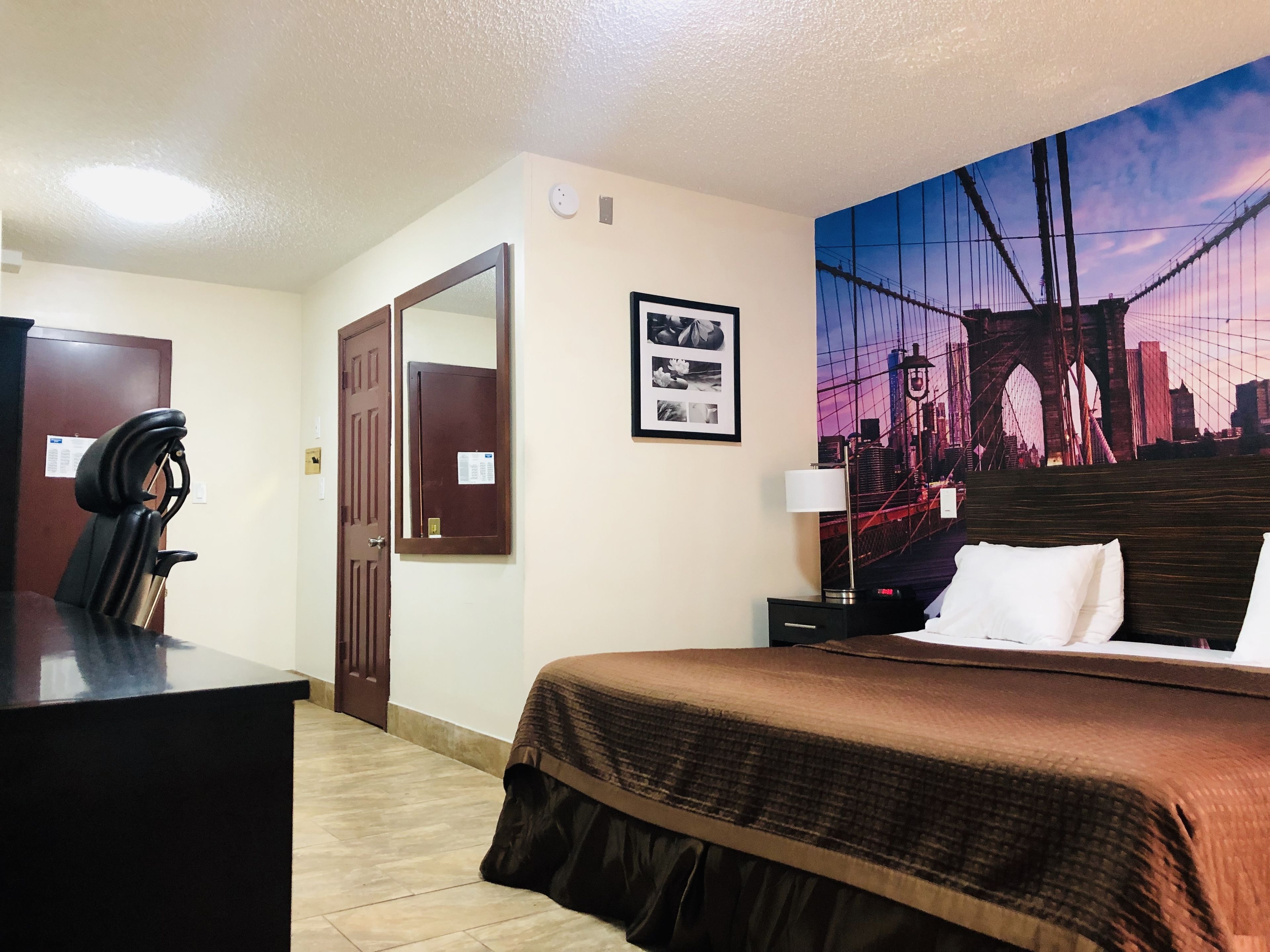 Une chambre d’hôtel avec un lit, un bureau et une chaise, une porte, une photo encadrée et un grand tableau représentant un paysage urbain accroché au mur.