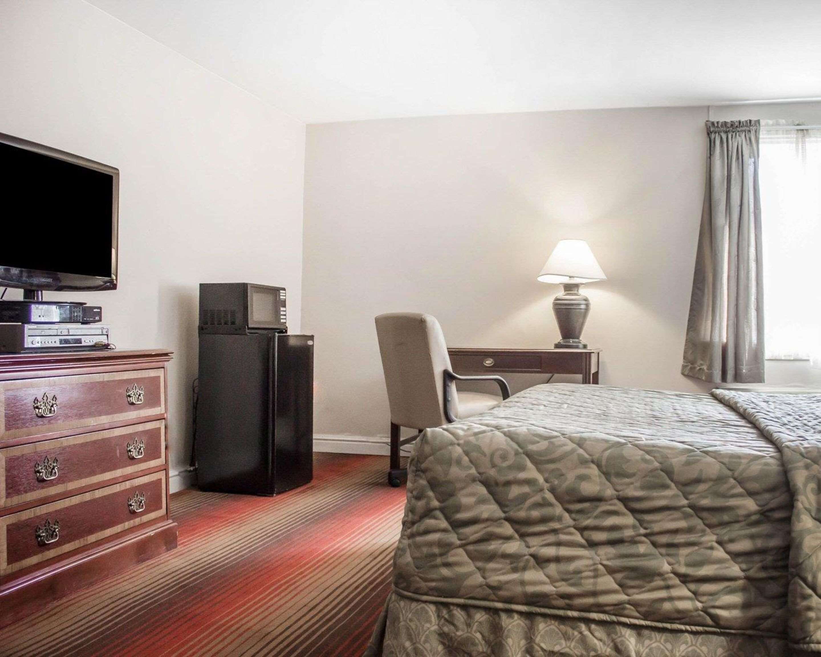 A hotel room with a bed, a desk with a lamp, a television on a dresser, and a chair.