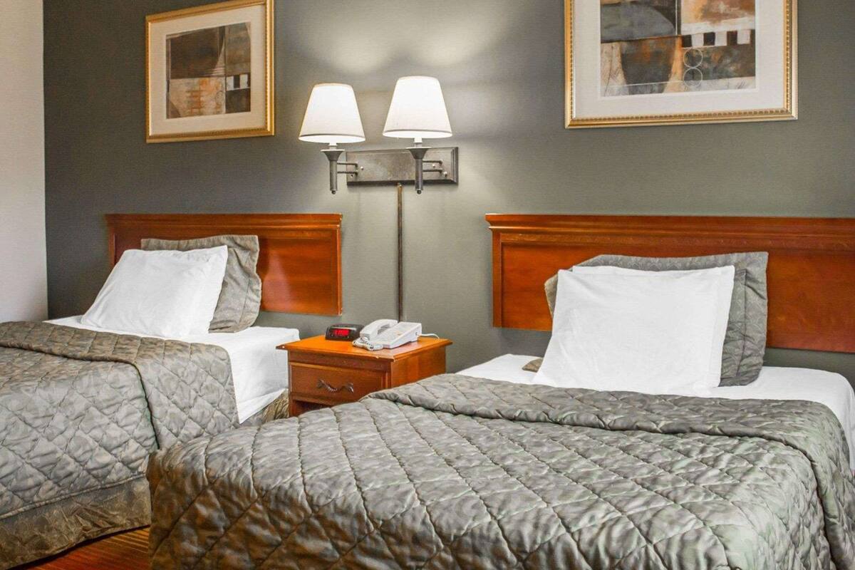 A hotel room with two beds, each with a quilted bedspread, a wooden headboard, a bedside table with a lamp, and a wall-mounted phone.