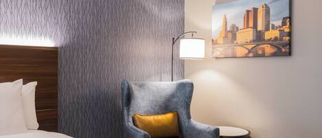 A hotel room with a bed, a grey armchair with a yellow cushion, a small round table, a lamp, and a cityscape painting on the wall.