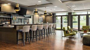 Un bar moderno con barra, varios taburetes y una zona de estar con sillas verdes y una mesa redonda.