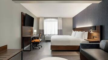 A modern hotel room with a large bed, a desk with a chair, a sofa, a TV mounted on the wall, and a window with curtains.