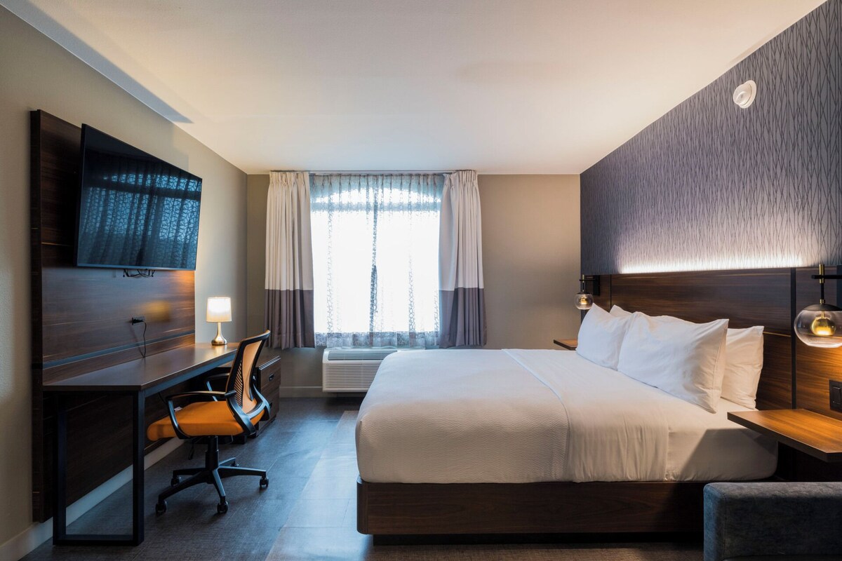A hotel room with a large bed, a desk with a chair, a TV mounted on the wall, and a window with curtains.