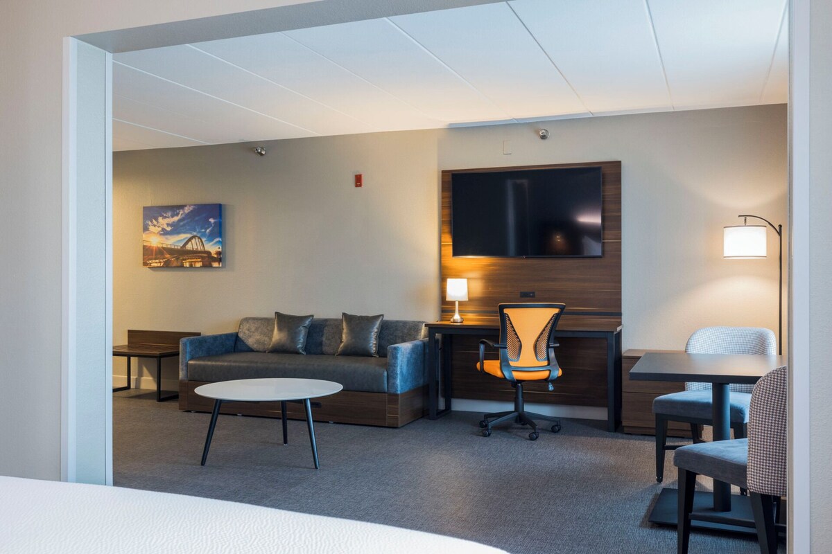A hotel room with a sofa, a small round table, a desk with a chair, a TV mounted on the wall, and a painting on the wall.