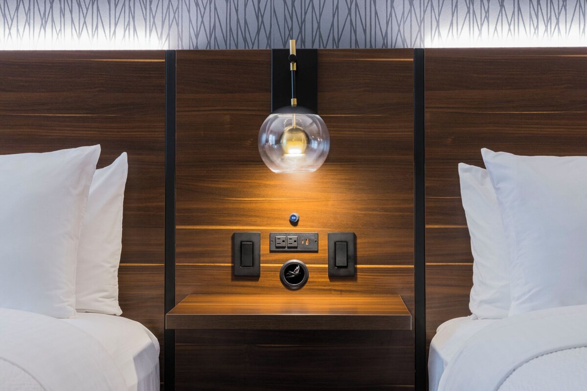 A hotel room with a wooden headboard, two white pillows, a bedside table with a lamp, and a control panel.