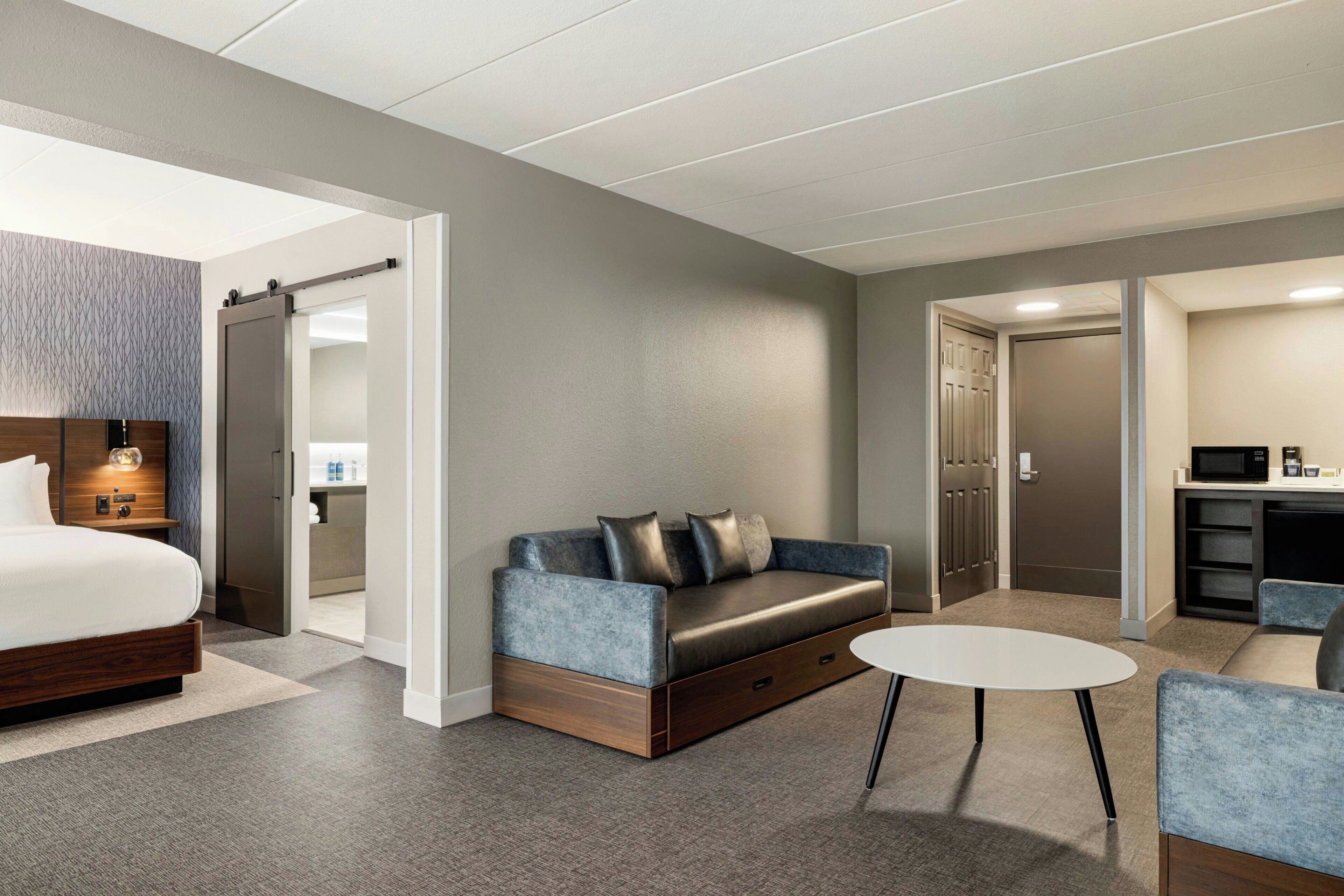 A modern hotel room with a bed, a sofa, a small table, and a kitchenette.