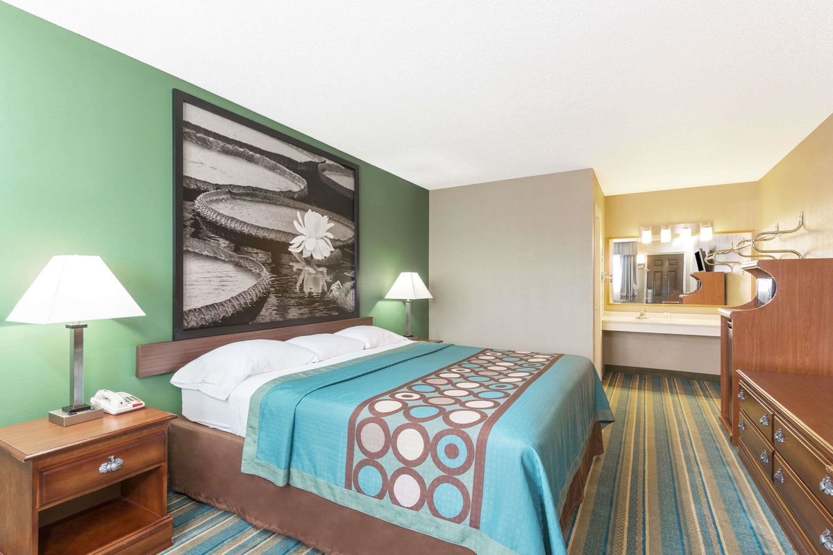 A hotel room with a bed, two bedside lamps, a telephone, a large painting, a mirror, and a wooden dresser.