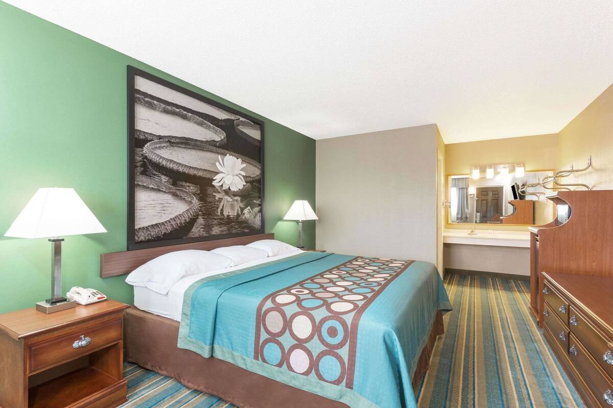 A hotel room with a bed, bedside lamps, a telephone, a large artwork on the wall, a bathroom with a mirror, and a wooden dresser.
