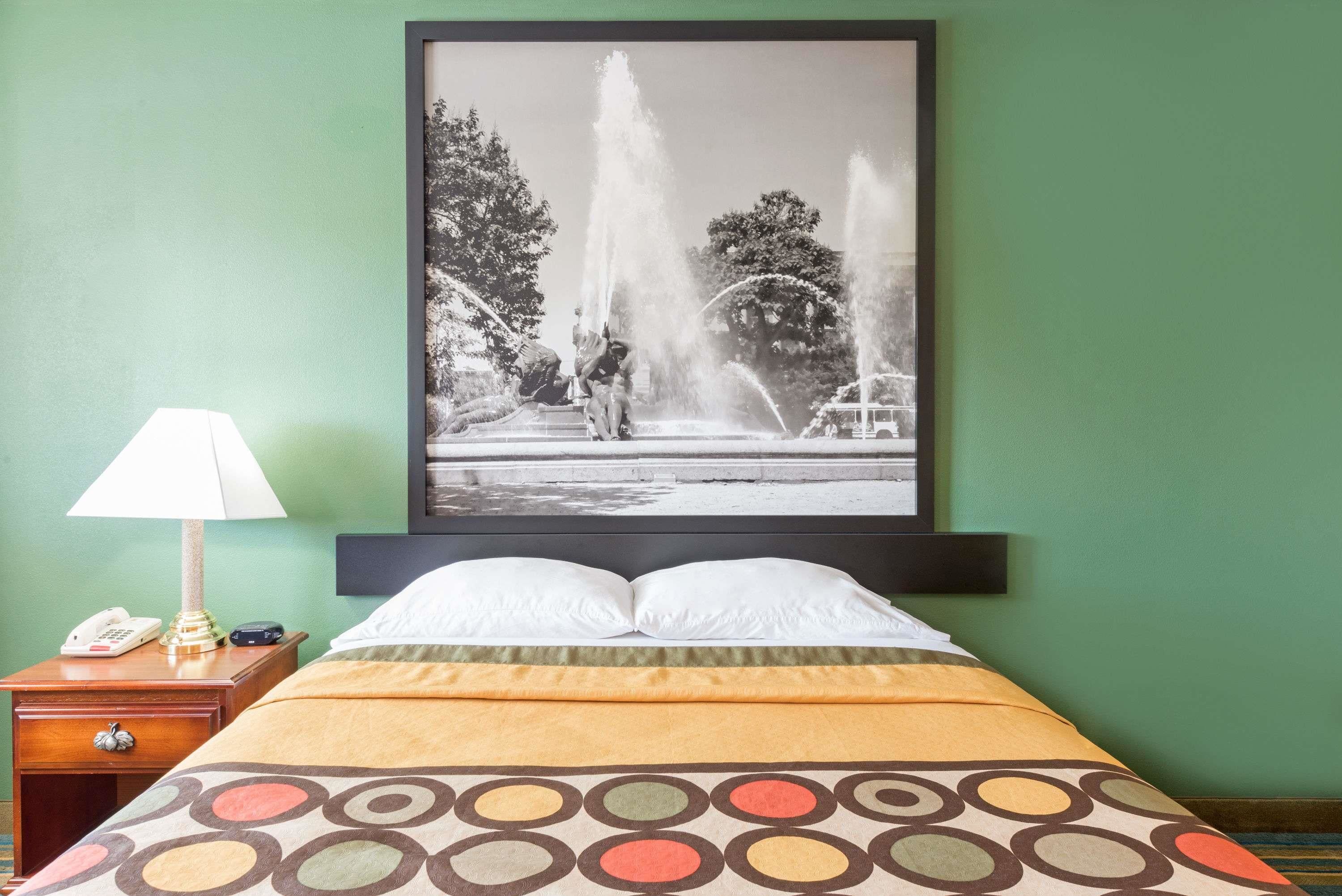 A hotel room with a bed, a nightstand with a lamp, a telephone, and a framed black and white photograph of a fountain.