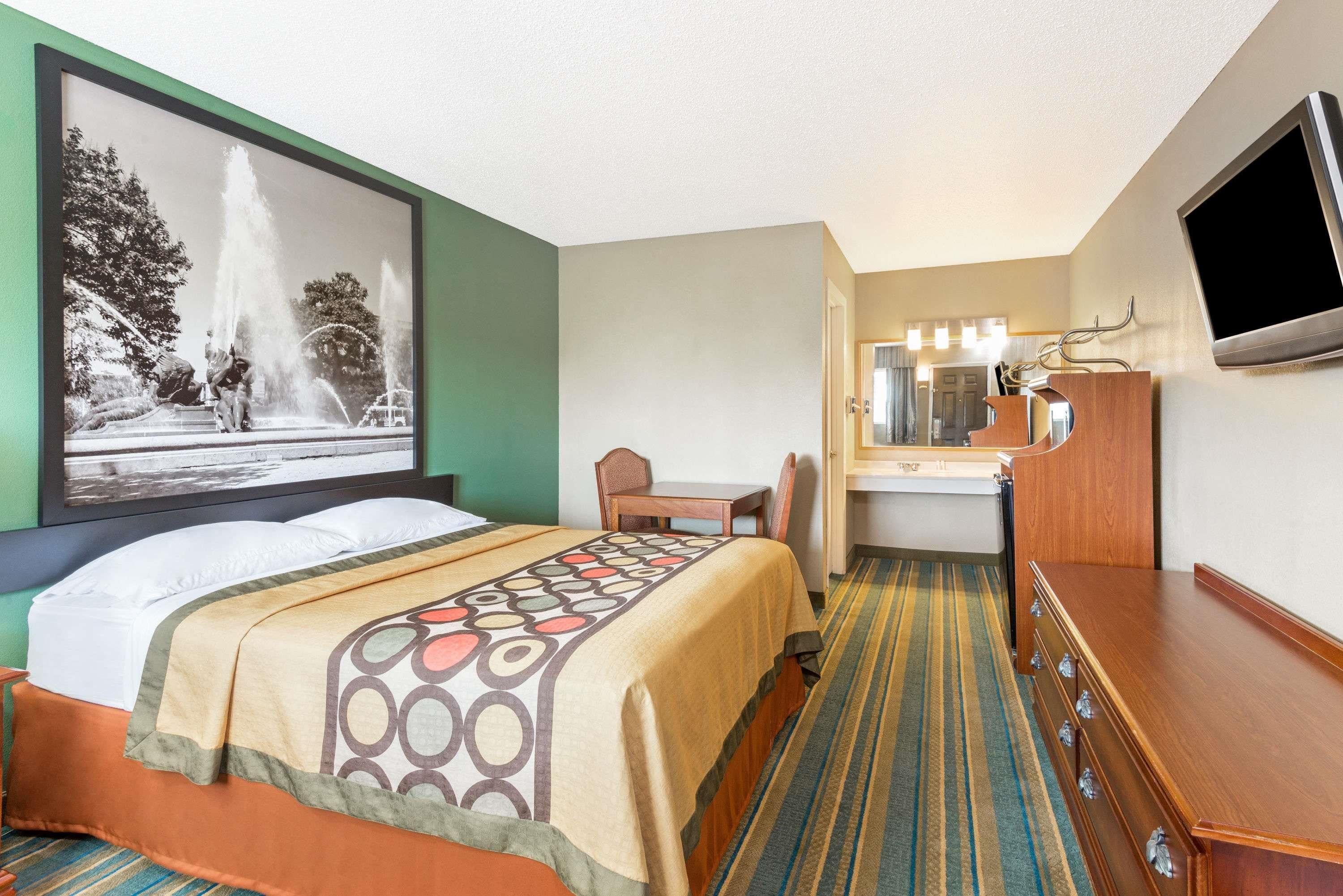 A hotel room with a bed, a desk, a chair, a TV, and a bathroom with a mirror and lights.