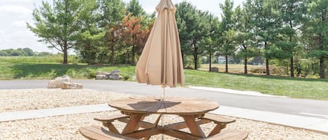 A wooden picnic table with an umbrella, surrounded by pebbles, in a park with trees and a road in the background.