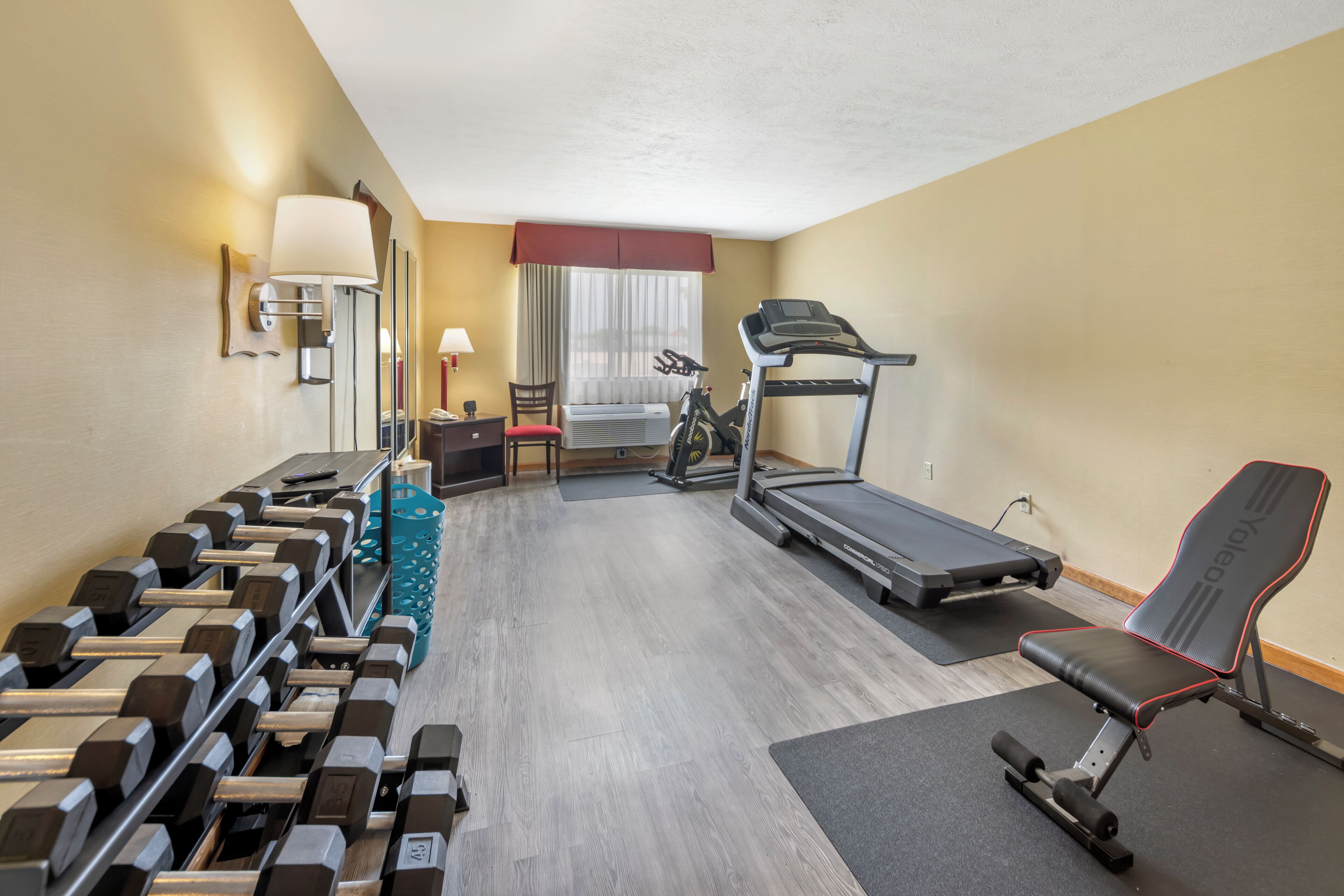 A hotel room with a treadmill, exercise bike, and a bench. There are dumbbells and a yoga mat on the floor.