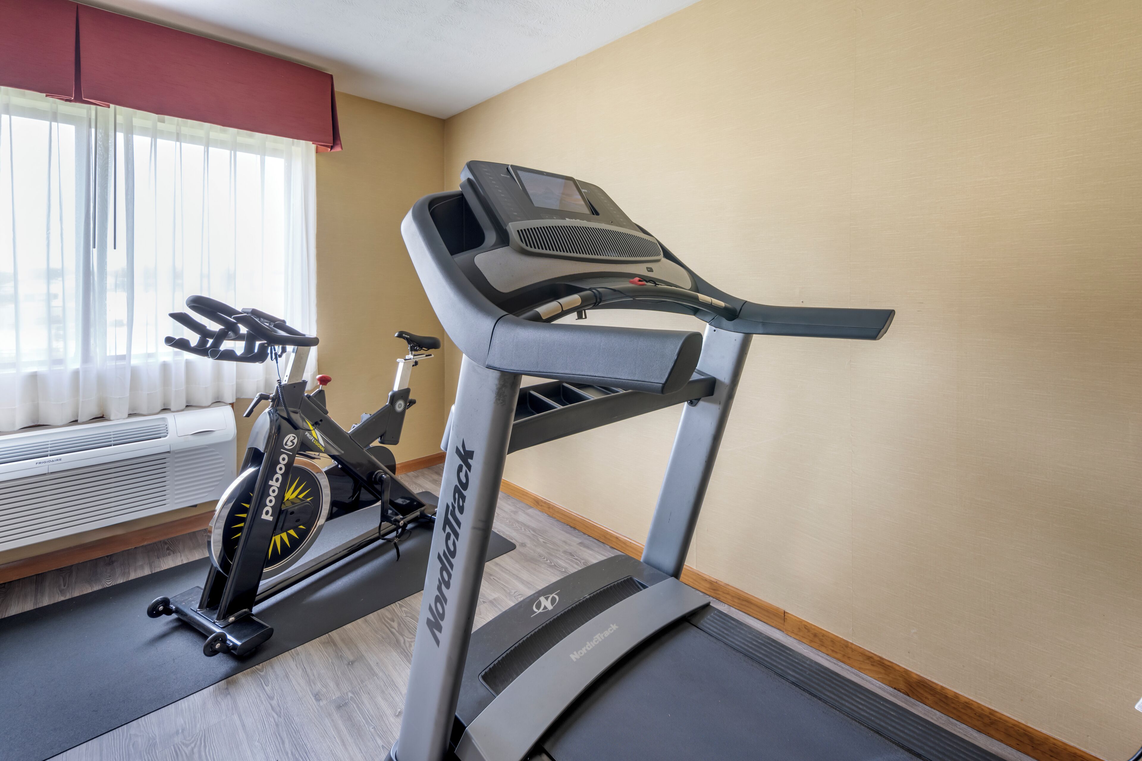Uma máquina de exercício NordicTrack e uma bicicleta ergométrica num quarto com janela, radiador e chão de madeira.