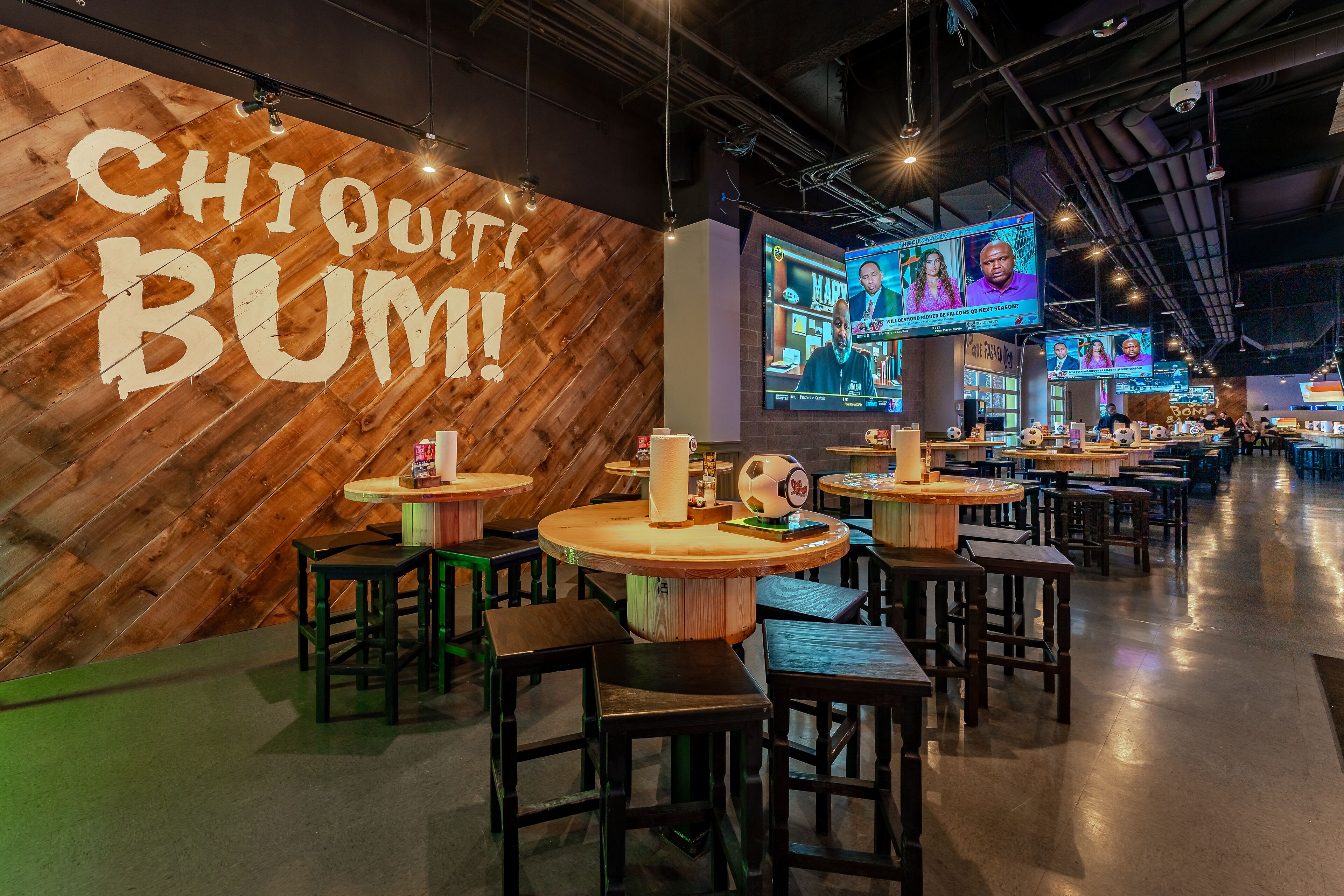 A restaurant with wooden walls and tables, a large screen displaying a show, and a sign that reads "CHIQUITI BUM!".