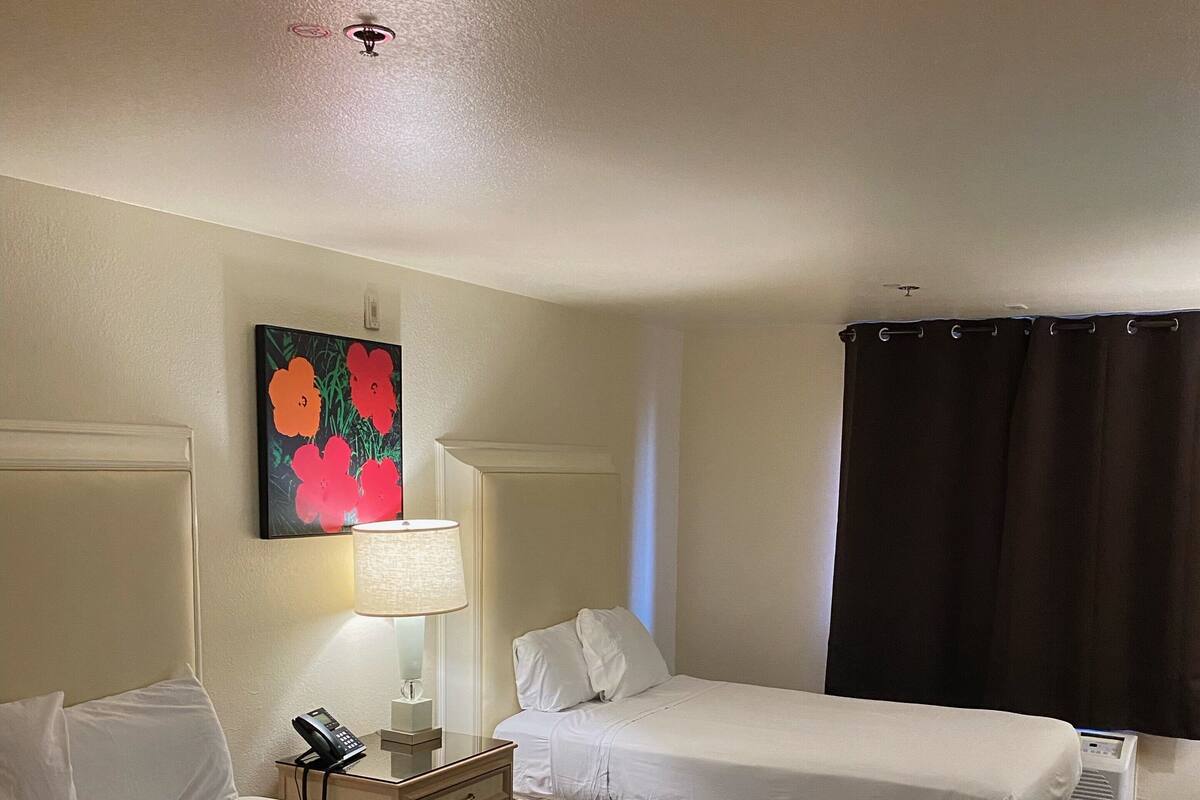 Une chambre d’hôtel avec deux lits, une table de chevet avec une lampe, un tableau au mur et un rideau foncé.