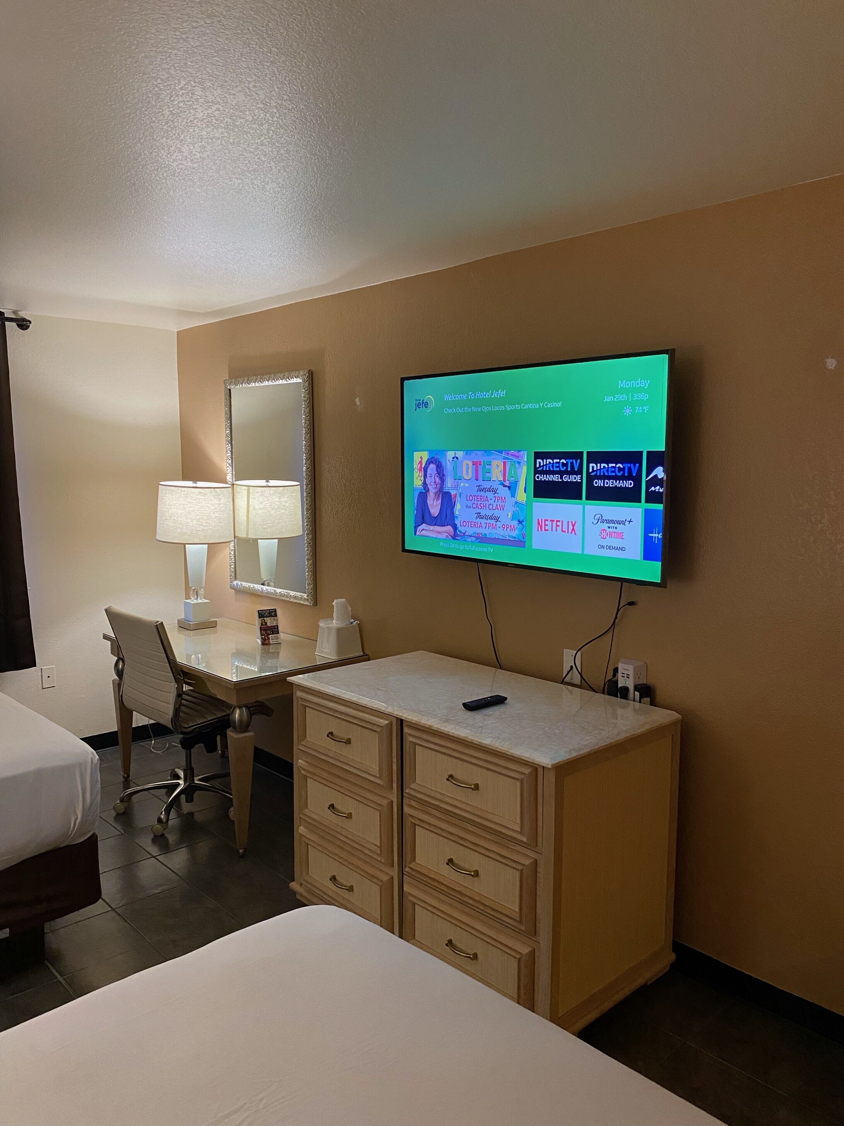 A hotel room with a bed, a desk, a mirror, a TV mounted on the wall displaying a welcome message, and a dresser.