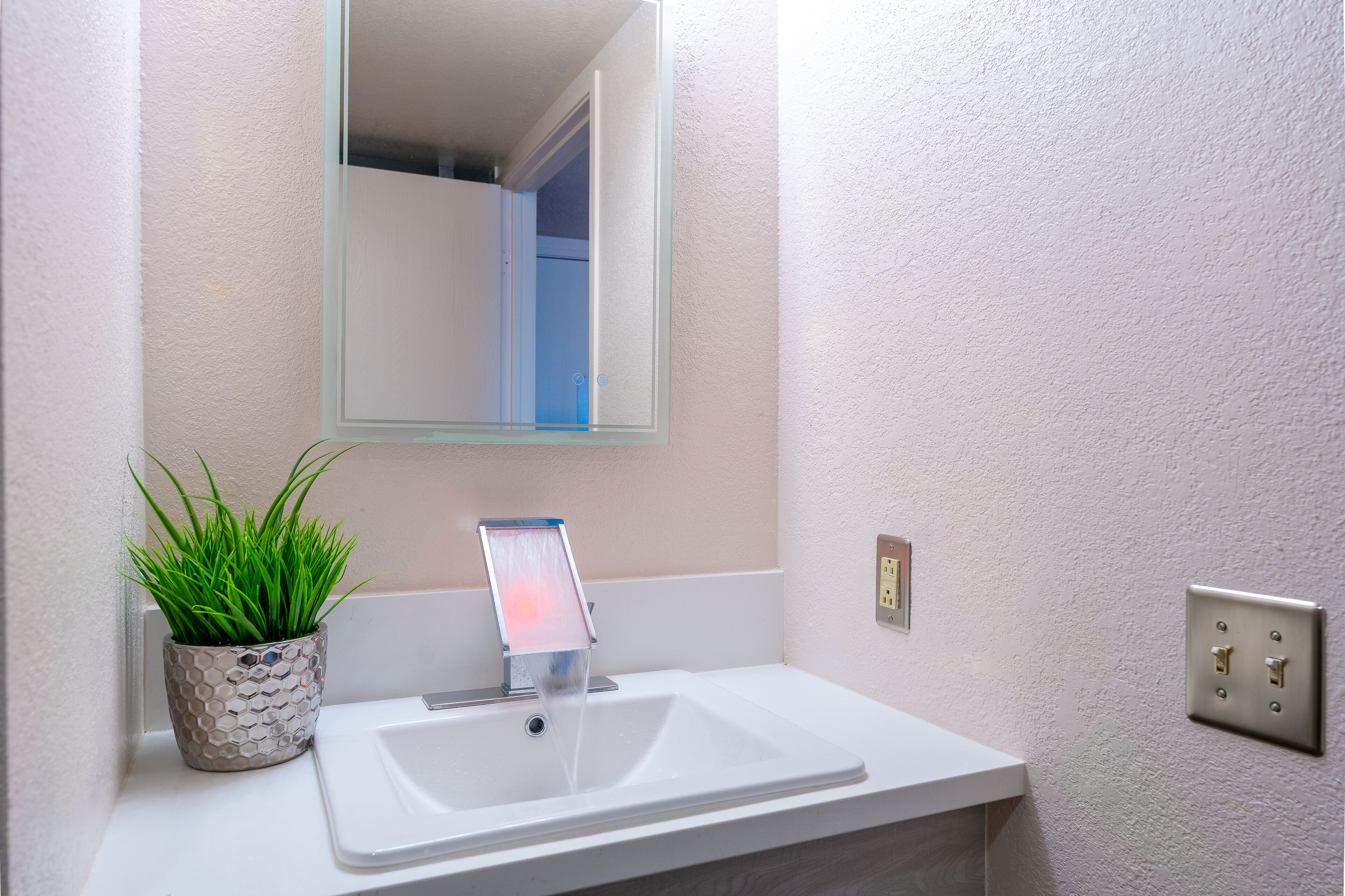 A bathroom with a white sink, a potted plant, a mirror, and a light fixture.