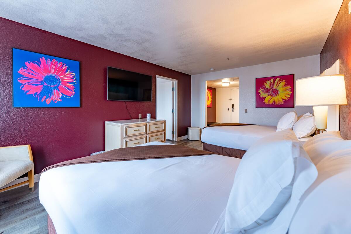 Une chambre d’hôtel avec deux lits, une commode, un téléviseur et deux grands tableaux représentant des motifs floraux.