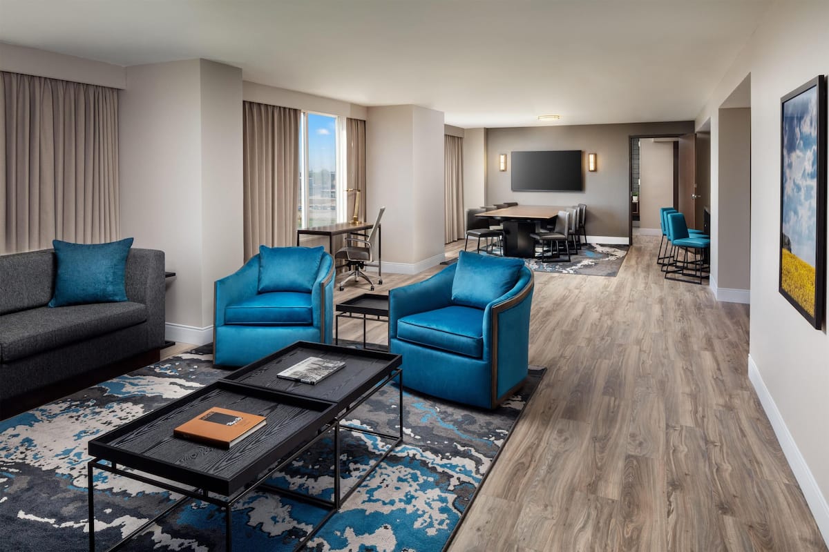 Ein modernes Hotelzimmer mit grauem Sofa, blauen Sessel, einem Couchtisch, einer Essecke mit Fernseher und einem großen Fenster mit Vorhängen.