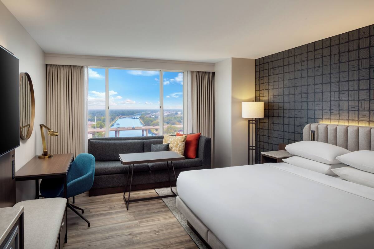 Ein Hotelzimmer mit einem großen Bett, einem Schreibtisch mit Stuhl, einer Sitzecke, einem Fernseher und Blick auf ein Gewässer und eine Brücke.