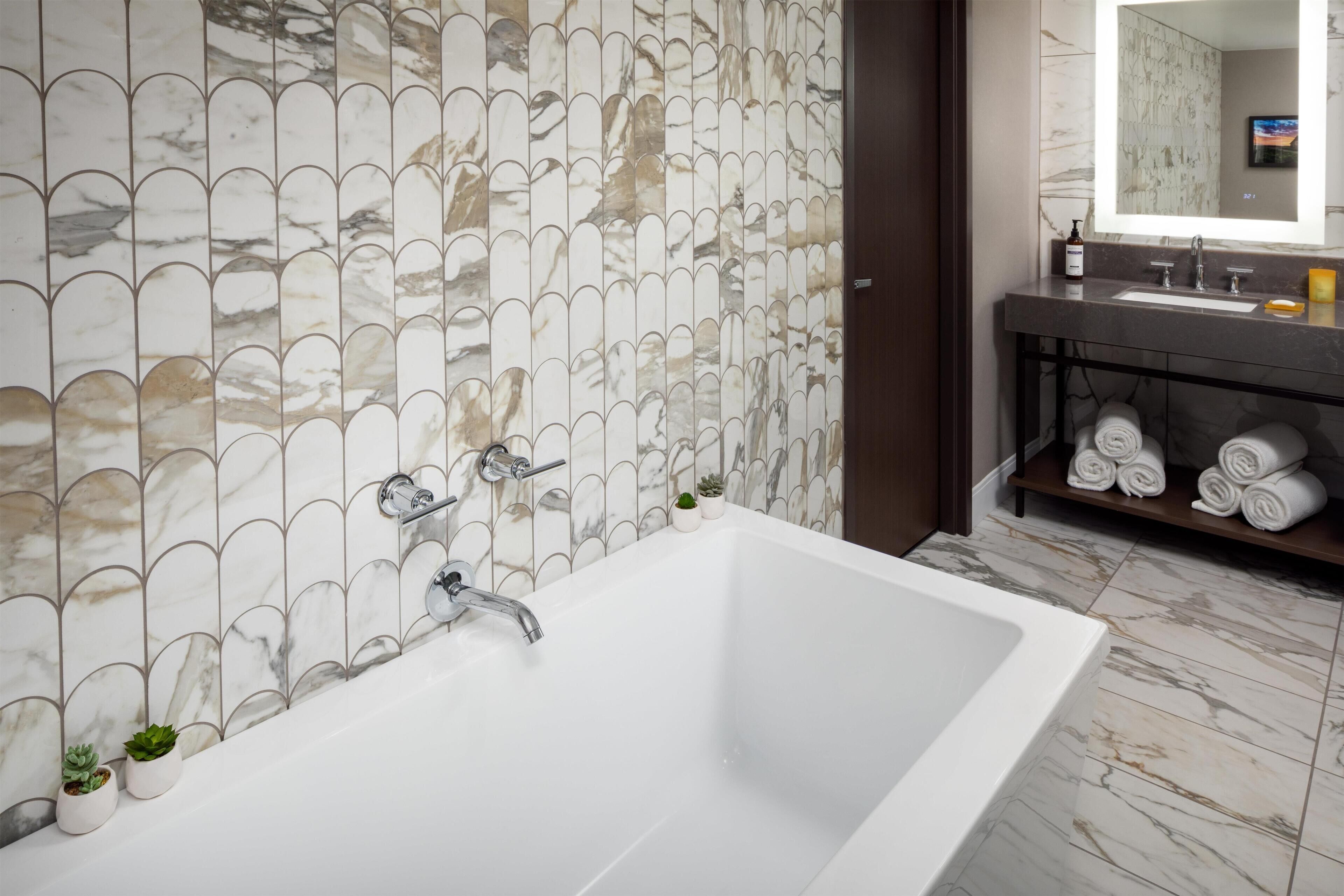 Une salle de bain moderne avec une grande baignoire, un mur aux motifs de marbre et une étagère en bois foncé où sont posées des serviettes.