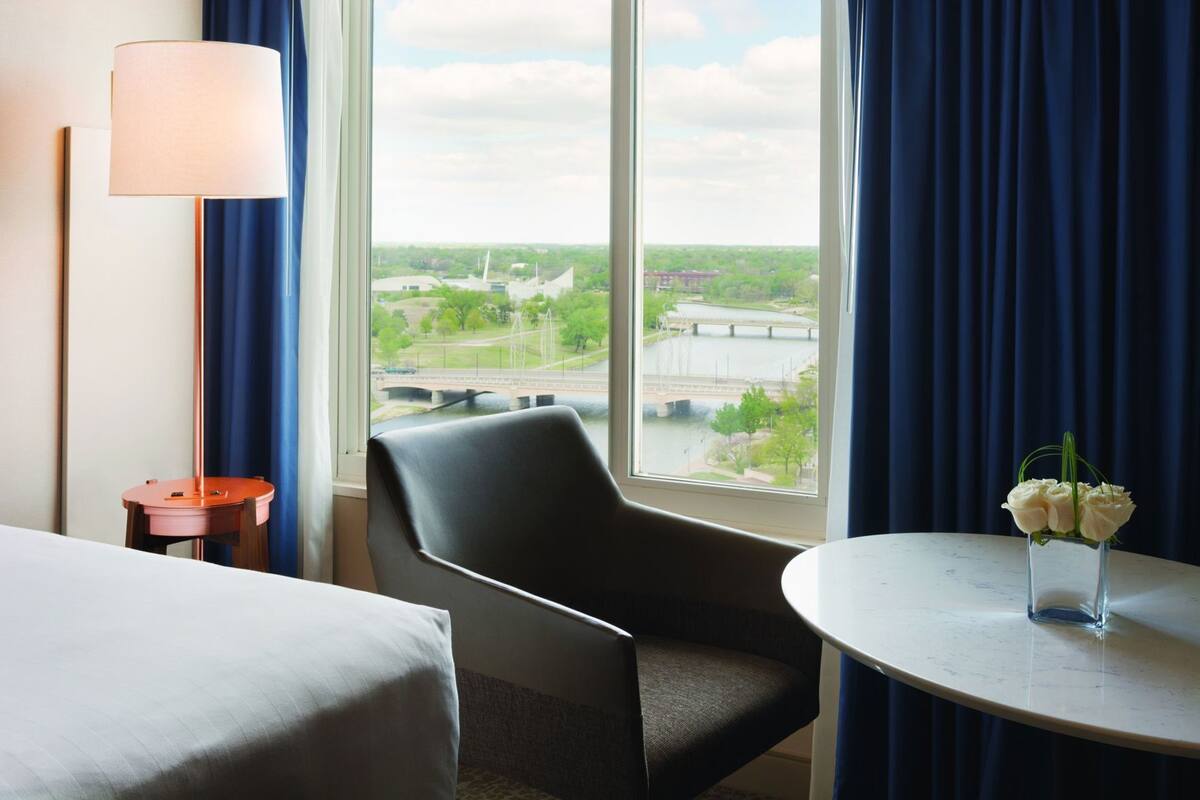 Ein Hotelzimmer mit einem Bett, einem Sessel, einem Tisch mit Blumenvase, einer Lampe und einem großen Fenster mit Blick auf eine Brücke und viel Grün.