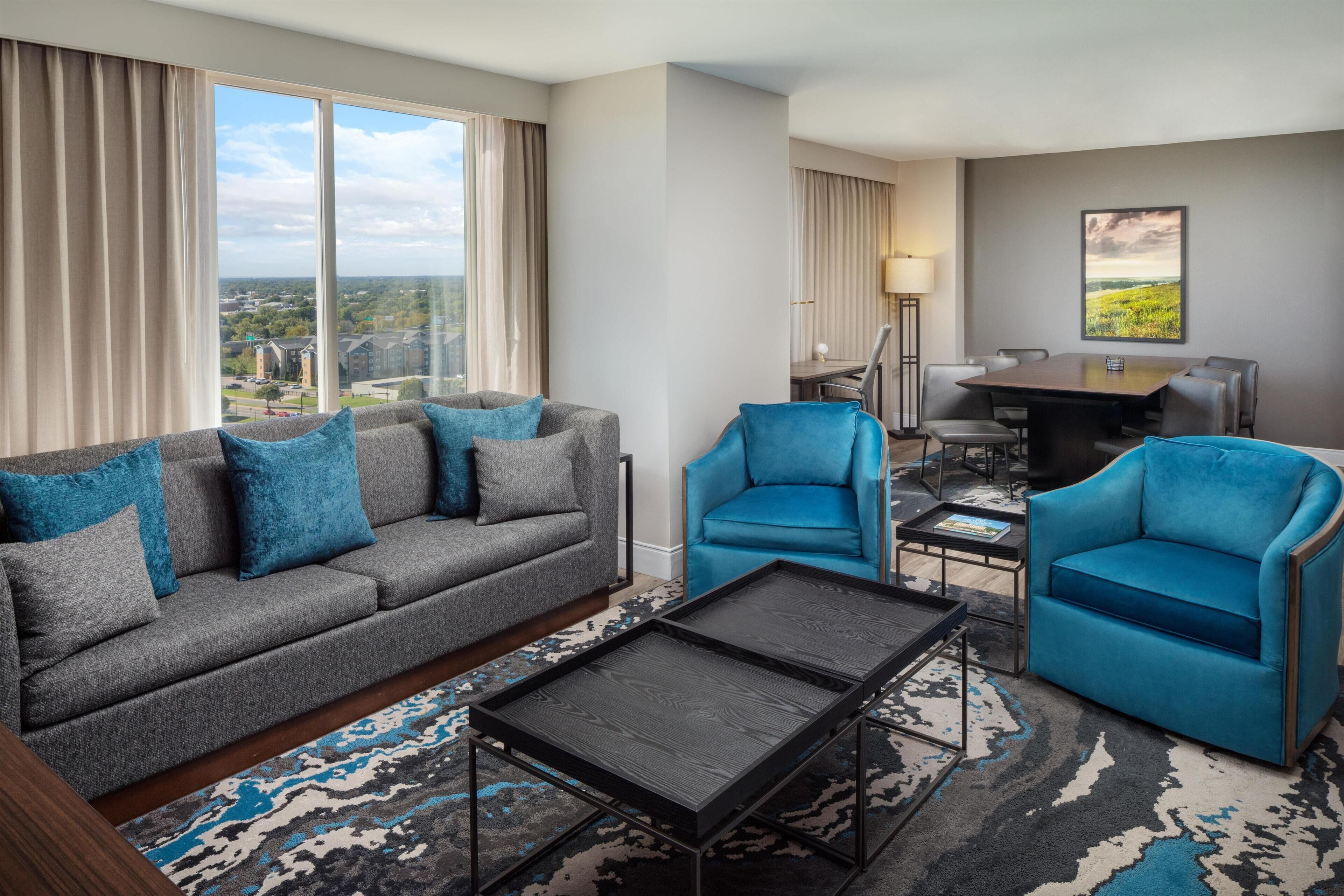 A modern hotel room with a gray sofa, blue armchairs, a small dining table, and a large window offering a city view.