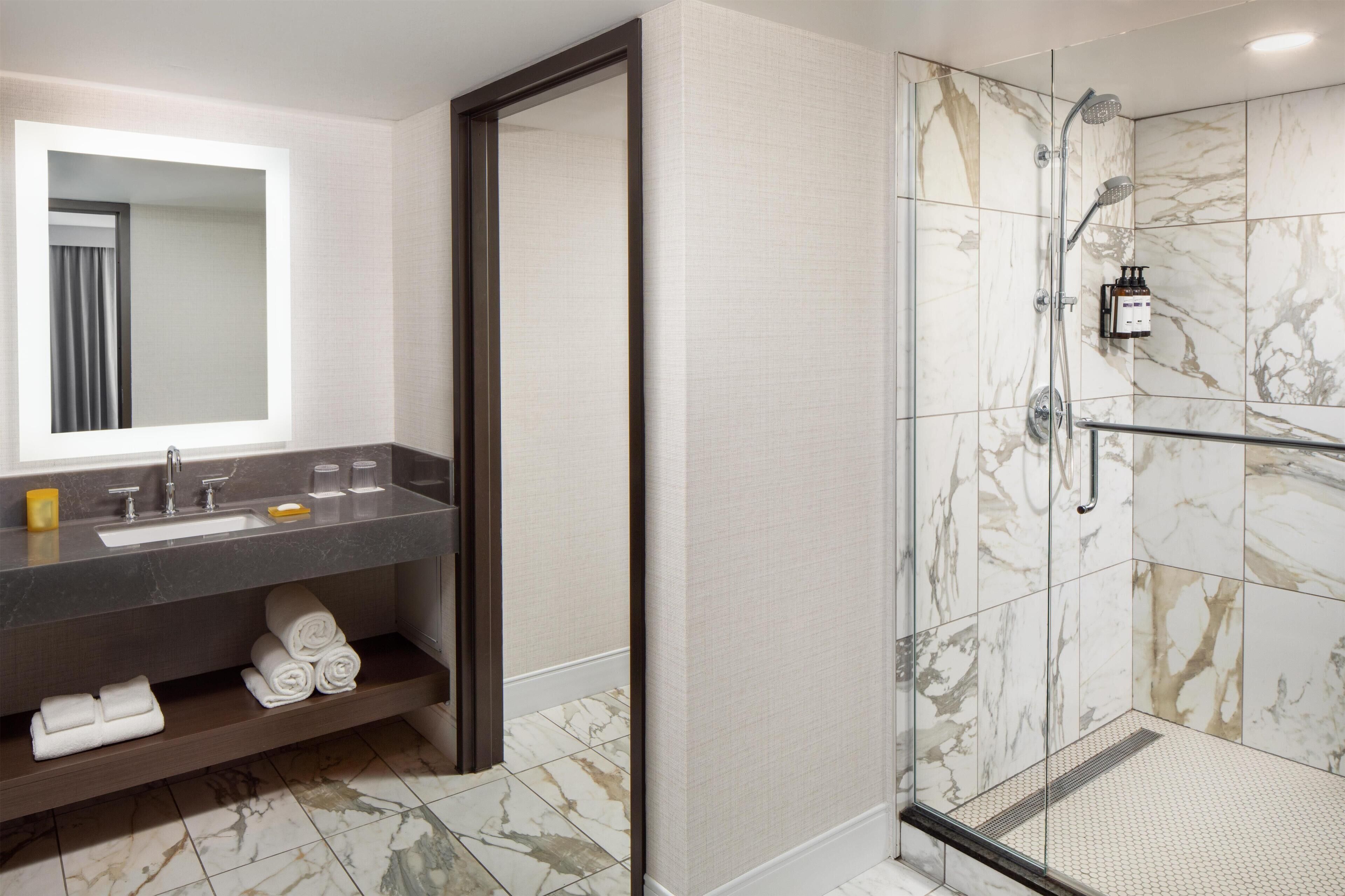 A modern bathroom with a marble shower, a large mirror, and a dark countertop with a sink and towel storage.