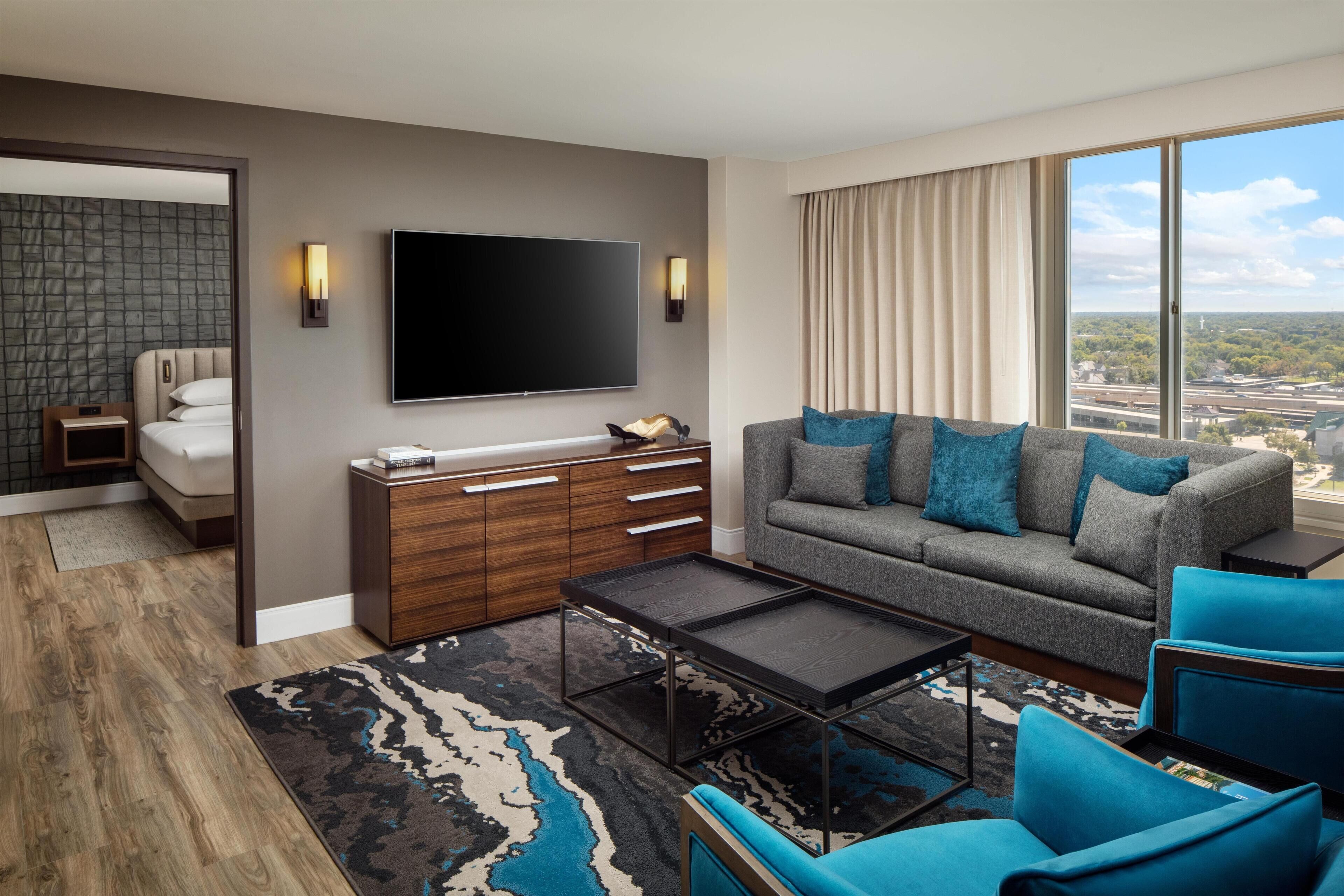 Habitación de hotel moderna con un televisor de pantalla plana grande, un sofá con cojines azules, un armario de madera y un ventanal con vistas a la ciudad.