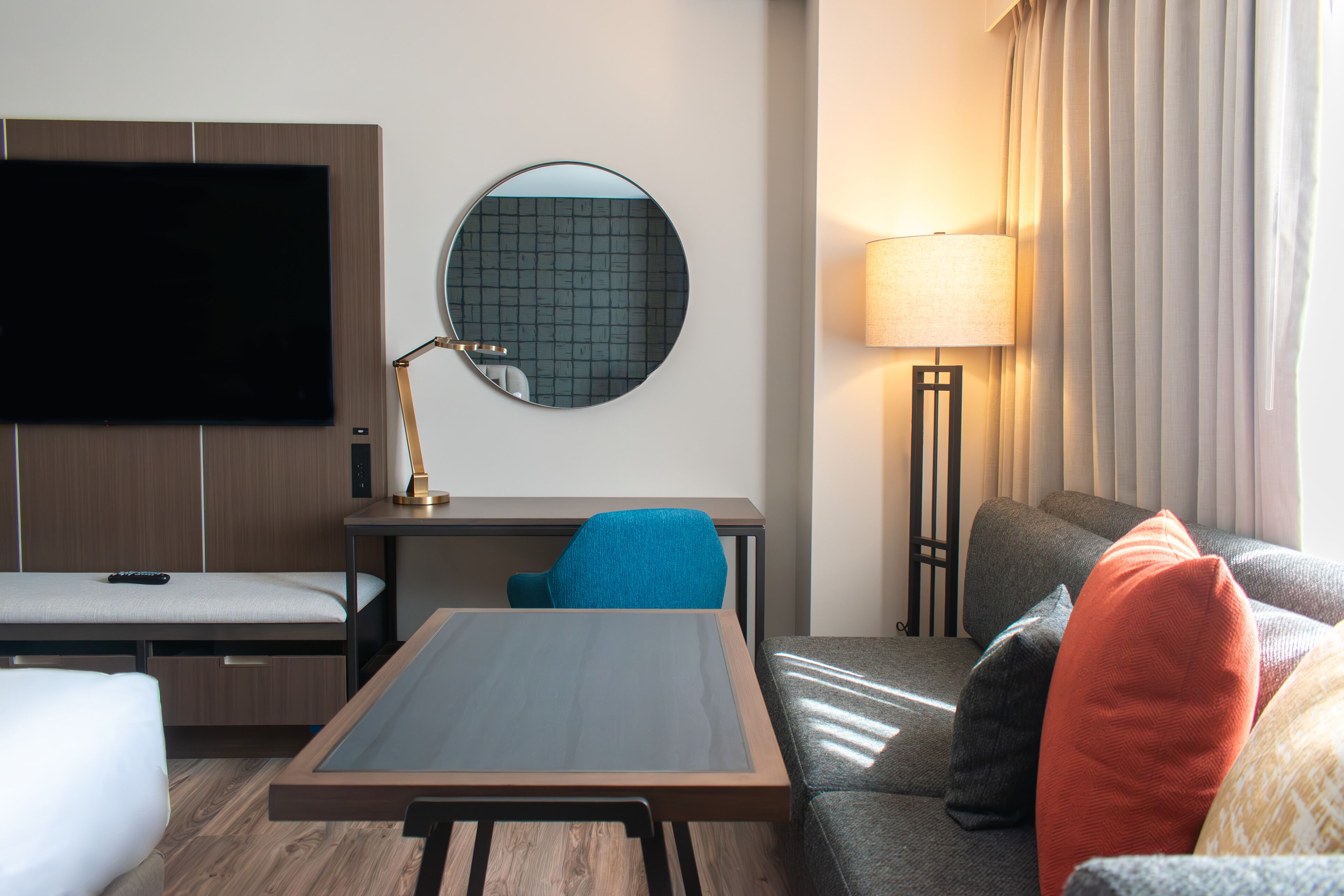 Habitación de hotel con televisión de pantalla plana, un espejo redondo, escritorio con lámpara, sofá con cojines y mesita de noche.