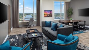 A modern hotel room with a large window offering a city view, a sofa with blue cushions, a dining table, and a flat-screen TV mounted on the wall. - Hyatt Regency Wichita (Wichita)