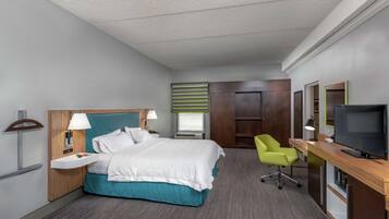 Habitación de hotel con cama, escritorio, silla, televisor y ventana con persianas.