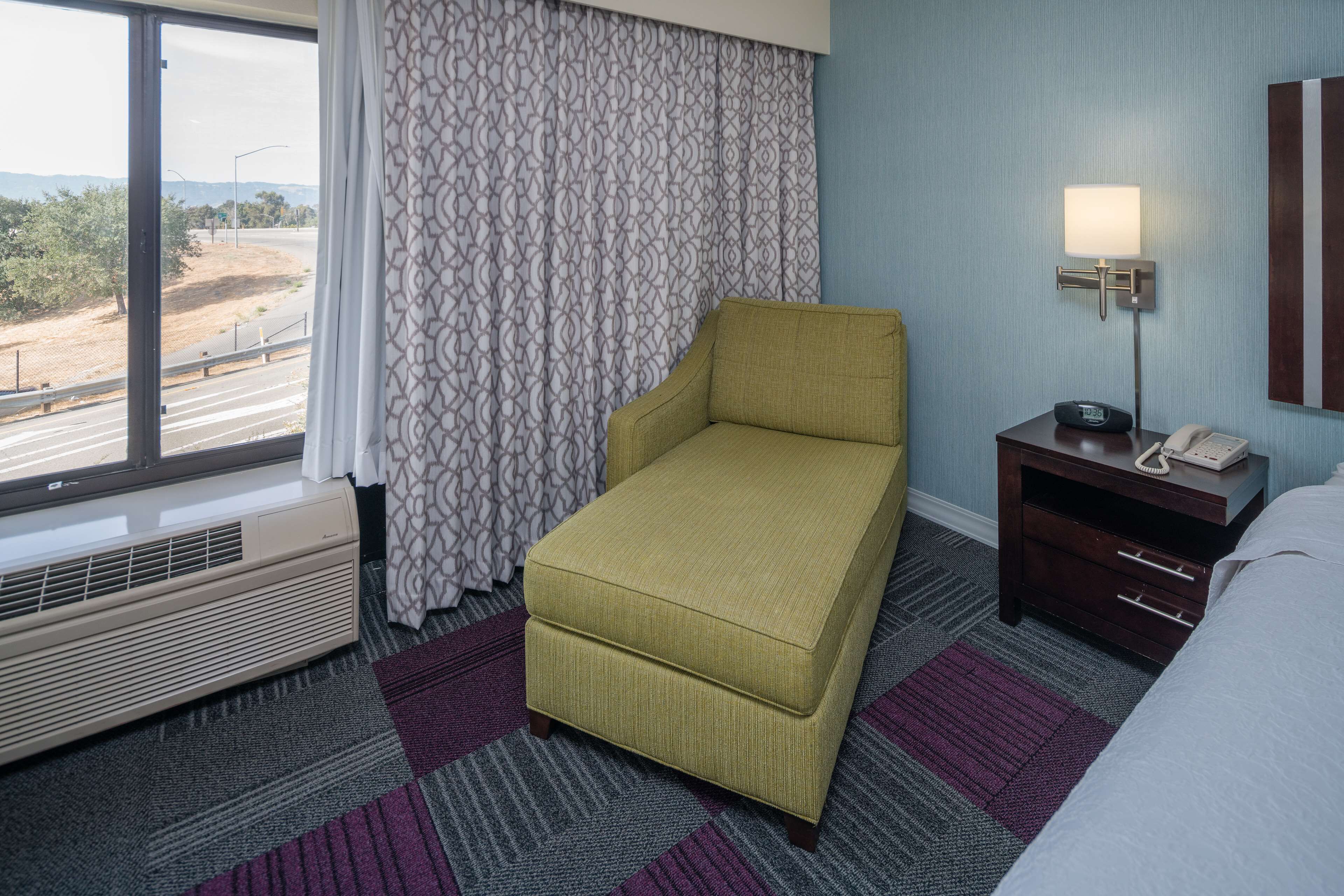 A hotel room with a single bed, a small sofa, a bedside table with a lamp, and a view of a road and trees through a window.